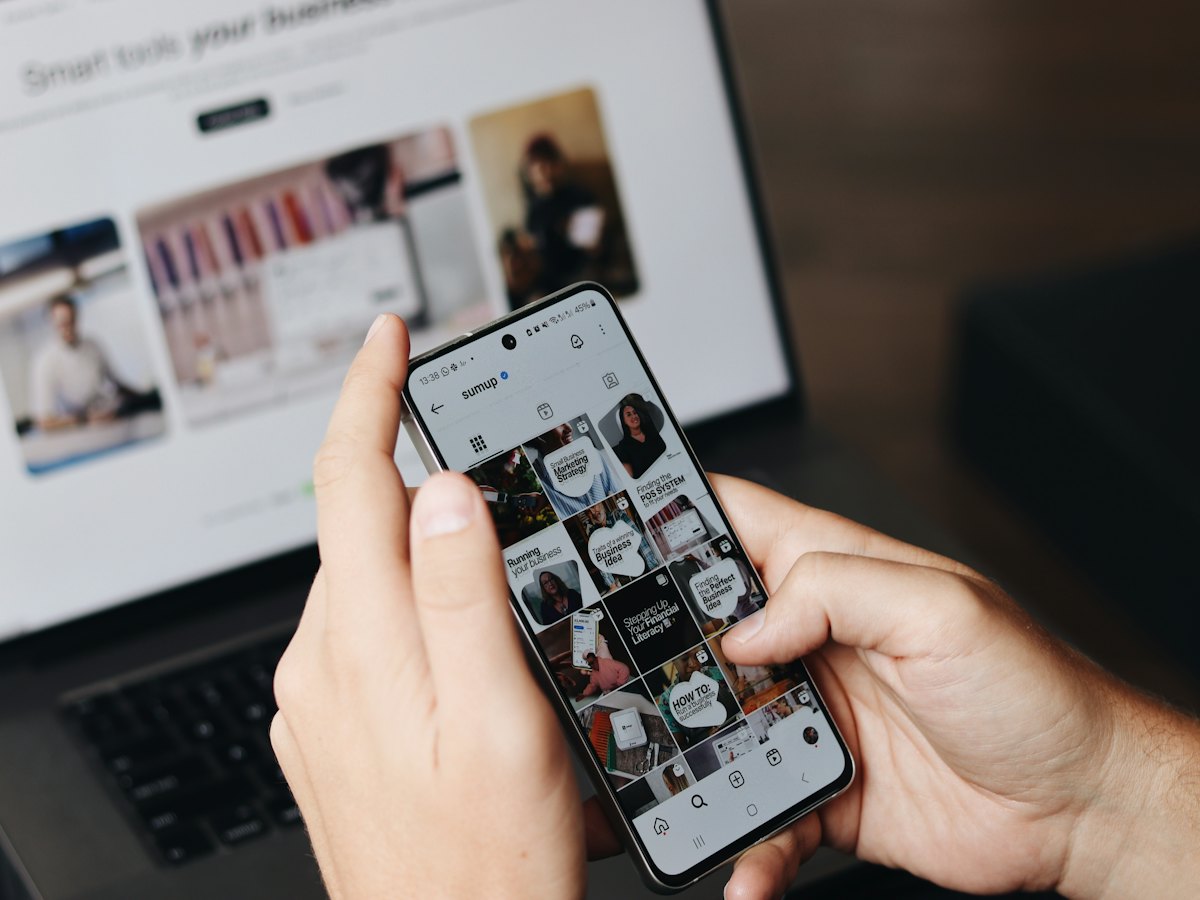 Person scrolling a social media feed on a phone in front of a laptop